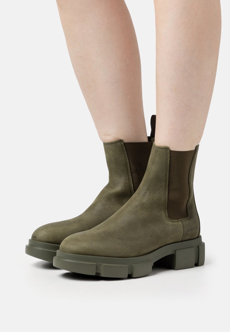 Bottines en cuir vert olive avec semelles épaisses portées sur des jambes nues sur fond blanc.