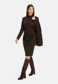 Braun gemustertes Kleid mit langen Ärmeln und Stehkragen. Das Modell trägt knielange, braune Wildlederstiefel. Insgesamt maßgeschneiderte und figurbetonte Gestaltung.