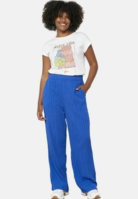 Bela majica s kratkimi rokavi z grafičnim dizajnom; modri široki hlače s pregibom. Manekenka pozira z rokami v žepih, obuta v superge.