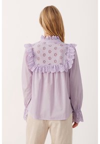 Blouse violet clair avec un col à volants et un motif ajouré, arborant des découpes florales et des poignets ajustés avec des détails similaires.