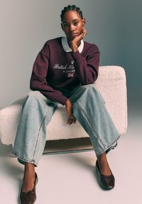 Burgundski pulover z belim ovratnikom, svetlo modri široki kavbojke in rjave balerinke. Model sedi na teksturirani beli kavču.