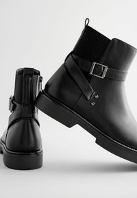Bottines noires en cuir avec des panneaux élastiques, des accents de boucles, une semelle plate et une tige en tissu texturé, présentées sous différents angles.