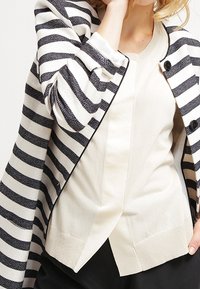 Randig kavaj i vitt och marinblått, med en texturerad tyg, öppen fram, och två svarta knappar. Bärs över en krämfärgad tröja.