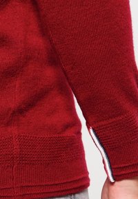 Pull en tricot rouge avec des poignets côtelés et des motifs texturés. Présente des rayures contrastantes en marine et blanc aux extrémités des manches. Texture douce.