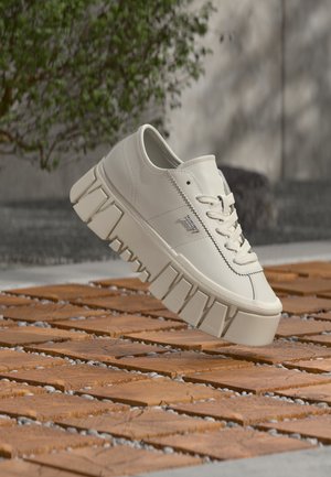 Baskets plateforme blanches avec semelle épaisse rainurée flottant au-dessus d’une surface en carreaux de bois en extérieur avec un fond de verdure flou.