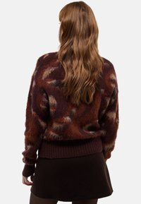 Un pull en tricot bordeaux confortable avec une finition douce et texturée, orné d'un motif abstrait et d'une taille côtelée, associé à une jupe sombre.