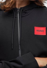 Close-up van een persoon in een zwart ritsjack met capuchon, met koordjes en een rood vierkant label met "HUGO" op de linker borst.