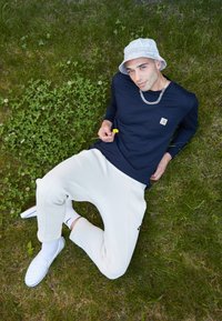 Tengerészeti hosszú ujjú póló zsebbekkel, krém színű jogger nadrág, fehér slip-on cipők és világosszürke konty kalap, fűre és lóherére helyezve.