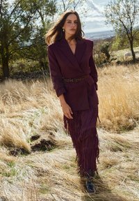 Bordowa marynarka o podwójnym zapięciu, zestawiona z frędzlowaną spódnicą. Noszona z wzorzystym paskiem i czarnymi botkami, na tle wysokiej trawy.