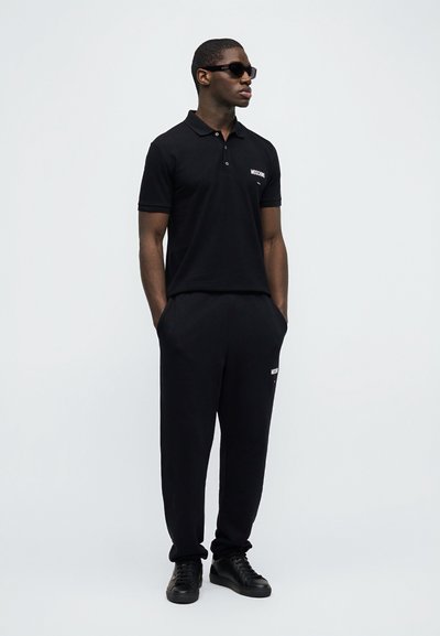 Άνδρας που φοράει μαύρο πόλο Moschino, μαύρο παντελόνι φόρμας, μαύρα αθλητικά παπούτσια και μαύρα γυαλιά ηλίου, στέκεται με τα χέρια στις τσέπες μπροστά σε λευκό φόντο.