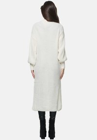 Cardigan lungo, a coste, di colore crema con maniche a palloncino, vestibilità comoda e lunghezza fino al polpaccio, abbinato a pantaloni neri in pelle aderenti e stivali.