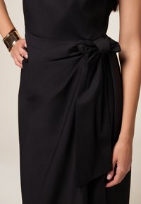 Zwarte jurk met een zijstrikdetail, gladde textuur en een getailleerde silhouet. Sluiting armband om de pols. Geen patronen of zichtbare hardware.