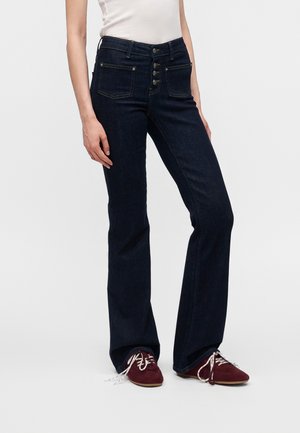 Kvinde iført mørkeblå viddejeans med knaplukning og vinrøde snørestøvler, stående mod en hvid baggrund.