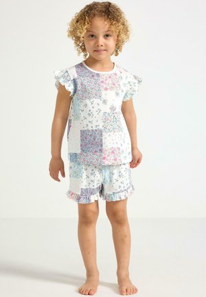 Lille barn med krøllet hår iført hvid blomstret patchwork top med flæseærmer og matchende shorts står barfodet på ensfarvet baggrund.