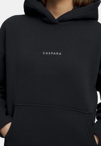 Svart hettegensere med en frontlomme og en liten hvit "CASPARA" logo. Laget av myk stoff med snor i hetten og ribbede mansjetter.