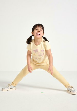 Gul bomuld t-shirt med sommerfugleprint, parret med ribbede gule leggings. Barnet har sølvfarvede sneakers med hvide såler på og står i en legesyg positur.