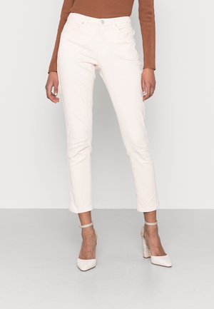 Femme portant un pantalon slim court écru, un haut marron à manches longues et des chaussures blanches à talons hauts avec des brides à la cheville, debout sur un fond uni.