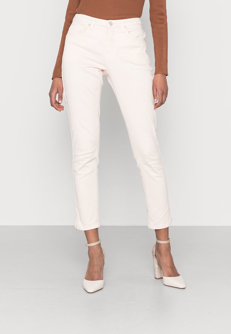 Femme portant un pantalon slim court écru, un haut marron à manches longues et des chaussures blanches à talons hauts avec des brides à la cheville, debout sur un fond uni.