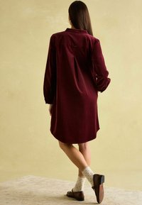 Bordeauxfarbenes, langärmliges Hemdkleid mit Kragen, ausgestattet mit einer Rückenfalte, geschwunghem Saum, getragen mit hellen, gepunkteten Socken und braunen Schuhen.