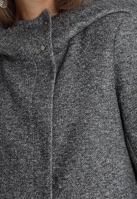 Šedá pletená bunda s texturovaným povrchem, vysokým límcem, zapínáním na druky a ležérním designem. Akcenty zvýrazňují hloubku látky.
