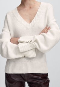 Pull en maille couleur crème avec col en V, manches oversize, poignets côtelés et boutons décoratifs. Texture lisse et motif minimal.