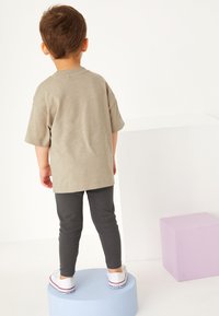 Kind trägt ein hellgraues T-Shirt und dunkelgraue Leggings. Das Shirt hat kurze Ärmel, und das Kind steht auf bunten Blöcken.