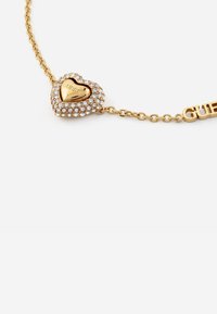 Bracciale in tono oro con un ciondolo a forma di cuore decorato con strass e un interno oro lucido, con una accentuazione del logo "GUESS" sulla catena.