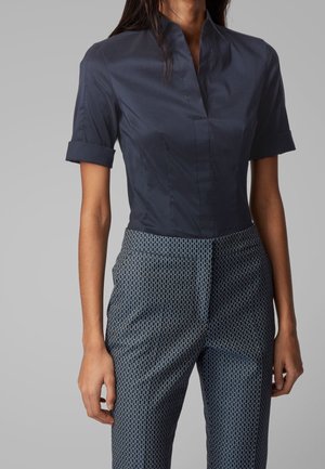 Femme portant un chemisier ajusté à manches courtes bleu marine avec des revers roulés et un pantalon taille haute à motifs bleu marine et blanc, posant devant un fond gris.