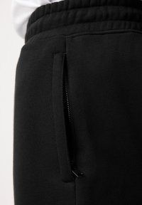 Gros plan d'un pantalon de survêtement noir avec une taille élastique et une poche verticale zippée sur le côté.