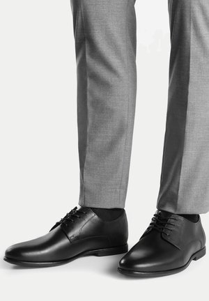 Czarne skórzane półbuty o gładkim wykończeniu, z ostrymi noskami i sznurowaniem na pięć dziurek, zestawione z szarymi, dopasowanymi spodniami.