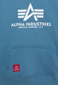 Felpa blu con logo bianco "ALPHA INDUSTRIES" e una tasca. Presenta un'etichetta rossa all'orlo. Tessuto morbido e vestibilità regolare.