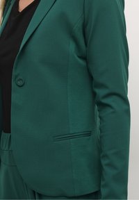 Blazer verde a medida en tejido texturizado, con cierre de un solo botón, un bolsillo discreto y corte ajustado, combinado con una blusa negra.