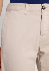 Beiga byxor med knappstängning, bältesöglor och detaljerad sömnad, bärs med en mörkblå topp synlig vid midjan.