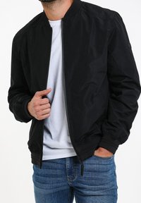 Veste bomber noire avec fermeture éclair à l'avant, poignets et col côtelés. Portée par-dessus une chemise blanche, et assortie à un jean en denim bleu.