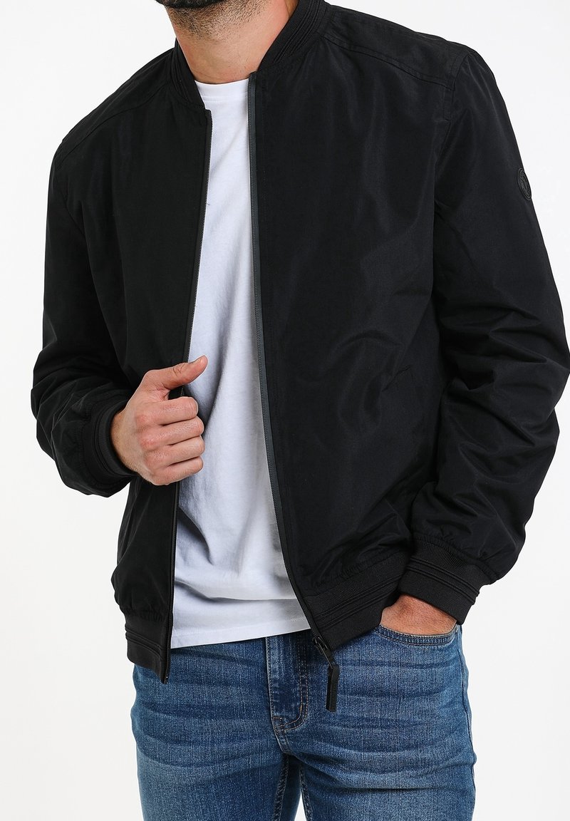 Veste bomber noire avec fermeture éclair à l'avant, poignets et col côtelés. Portée par-dessus une chemise blanche, et assortie à un jean en denim bleu.