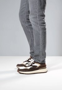 Braun-weiße Turnschuhe aus Wildleder und Mesh-Material mit sportlichem Design, dicker Sohle, gepolstertem Zungenbereich und kontrastierenden Akzenten. Graue Jeans sichtbar.