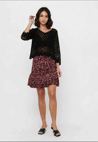 Suéter cropped de malha aberta preto com decote em V, combinado com uma saia com padrão floral em tons de preto e vermelho com folhos. Sandálias slide pretas.