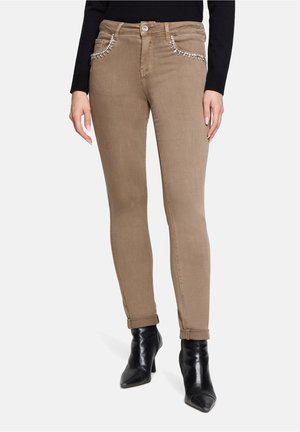 Woman wearing beige slim-fit jeans with jeweled front pockets, rolled cuffs, black pointed-toe ankle boots, and a black long-sleeve top.