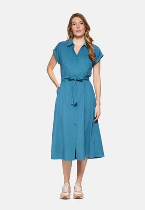 Mujer con un vestido midi abotonado en color verde azulado, con cinturón anudado, mangas cortas y zapatillas beige, de pie con una mano en el bolsillo.