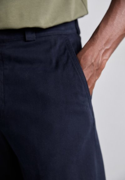 Marina bomullbyxor med en slät textur, sidofickor och en slim fit-design, tillsammans med en olivgrön skjorta.