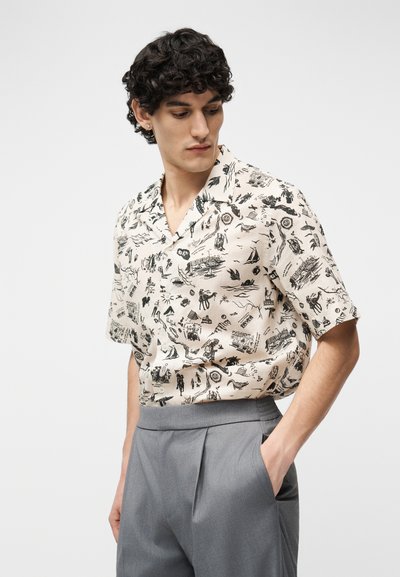 Νεαρός άνδρας με σγουρά μαλλιά που φοράει μπεζ πουκάμισο με κοντά μανίκια και μαύρα αφηρημένα σχέδια, καθώς και γκρι πλισέ παντελόνι, με το χέρι στην τσέπη.