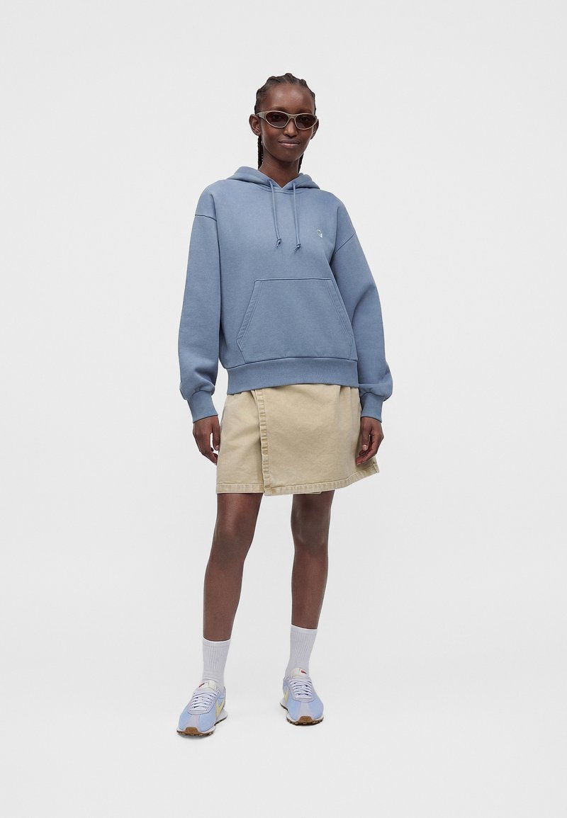 Person wearing blue hoodie, beige skirt, white socks, light blue sneakers, and dark sunglasses standing against plain background.