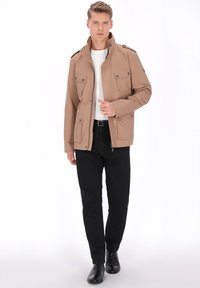 Tanhvid letvægtsjakke med lynlås og fire forlommer, båret over en hvid skjorte og kombineret med sorte bukser og støvler.