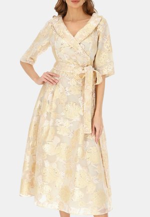 Kobieta w beżowo-złotej sukience kopertowej z kwiatowego brokatu, z rękawami do łokcia i kokardą wiązaną w talii.