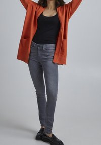 Oranžový pletený cardigan s kapsami, černý top, šedé úzké džíny a černé mokasíny, postava s rukama zdviženýma na světlém pozadí.