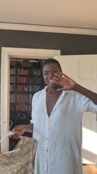 Jeune femme en chemise blanche souriante et faisant signe de la main, tenant une robe à motifs sur un cintre dans une pièce avec des étagères à livres en arrière-plan.