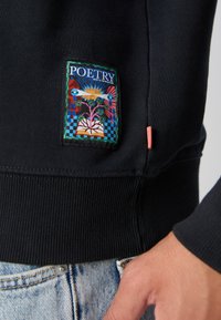 Mornarska dukserica s rebrastim rubom i manžetama. Sadrži šareno "POETRY" naljepnicu koja prikazuje stilizirane cvjetove i geometrijske oblike.