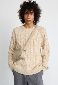 Pletena bež jopica z vzorcem kabel, v kombinaciji s sivimi prugastimi hlačami in majhno svetlo bež torbico preko telesa.