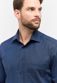 Uomo con capelli corti e castani e barba, che indossa una camicia button-up blu scuro testurizzata, che guarda a destra su uno sfondo bianco.