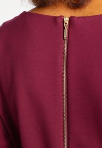 Lähikuva kullankeltaisesta vetoketjusta burgundin värisessä kankaan vaatteessa, jota henkilö käyttää.